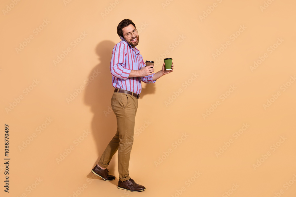 Fototapeta premium Full size photo of nice young male walk phone call hold coffee wear trendy striped formalwear isolated on beige color background