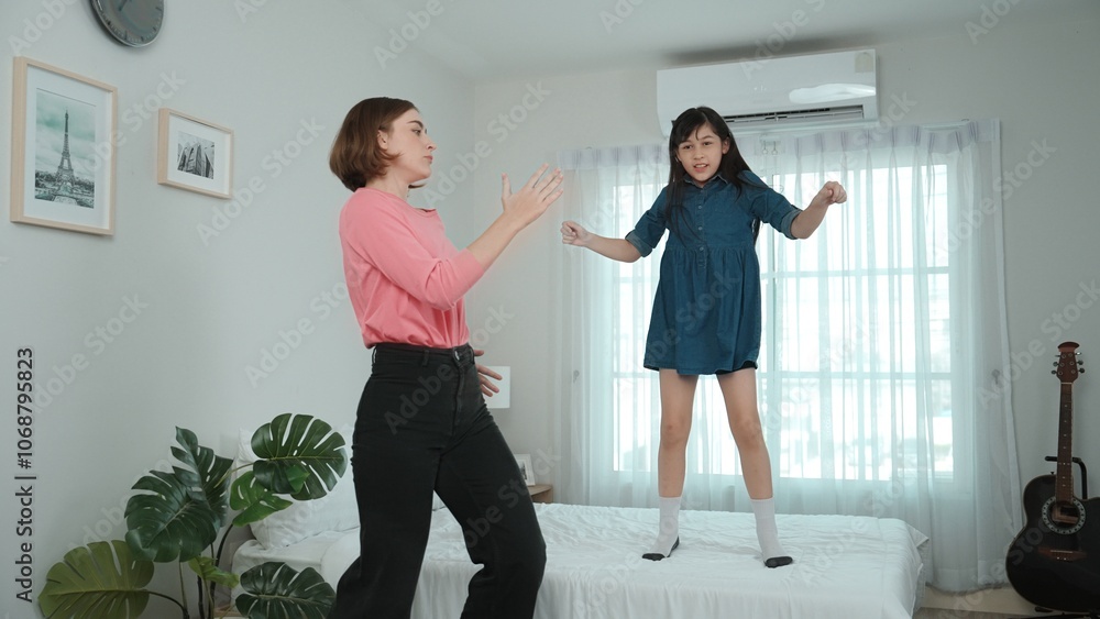 Fototapeta premium Happy attractive mother dancing to music together with asian daughter at bedroom. Caucasian mom dance or exercise while cute child move to music while jump to bed. Family recreation concept. Pedagogy.