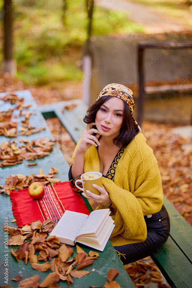 the beautiful brunette with a scarf on her head and a blanket is sitting at a table with a book, a cup and an apple in the autumn landscape. the beautiful brunette with a scarf on her head and a blanket is sitting at a table with a book, a cup and an apple in the autumn landscape.