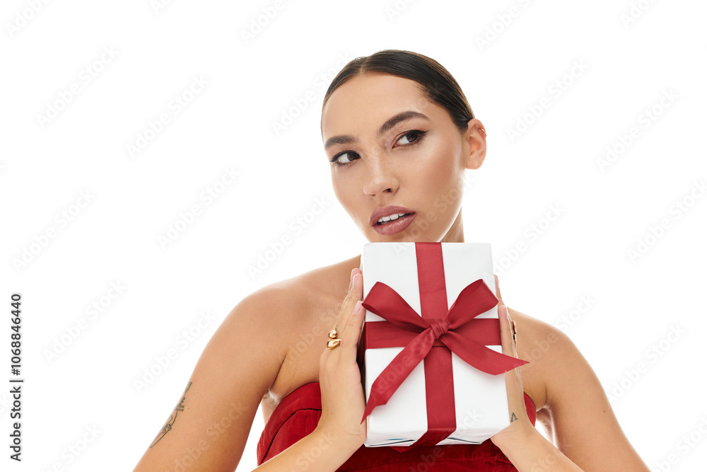 The woman, dressed elegantly, playfully poses while holding a gift with a red ribbon. The woman, dressed elegantly, playfully poses while holding a gift with a red ribbon.