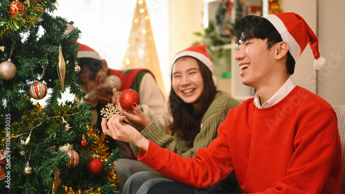 Three friends in Santa hats laugh and hold ornaments as they decorate the Christmas tree, sharing a joyful and playful moment in a warm holiday setting, Laughing Together on Christmas Day