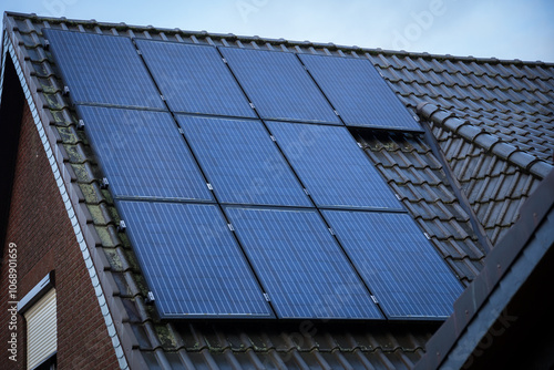 A slanted rooftop with solar panels installed, generating renewable energy. The panels are aligned on a tiled roof under a cloudy sky.