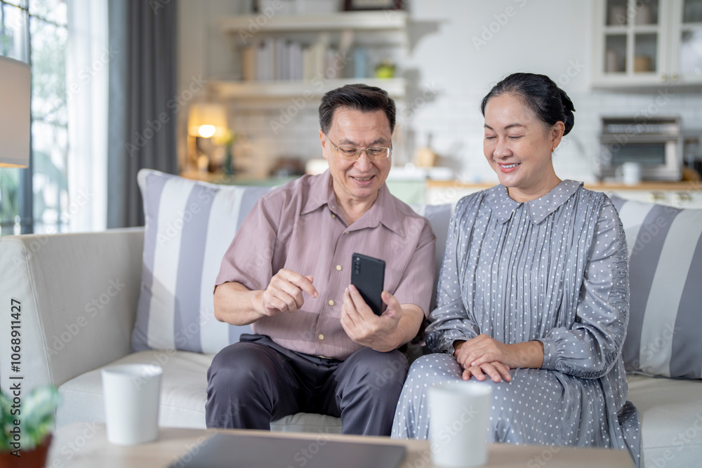 An elderly couple sits together in their cozy living room, smiling and enjoying a moment as they look at something on a smartphone. Their warmth and affection capture a peaceful home atmosphere. An elderly couple sits together in their cozy living room, smiling and enjoying a moment as they look at something on a smartphone. Their warmth and affection capture a peaceful home atmosphere.