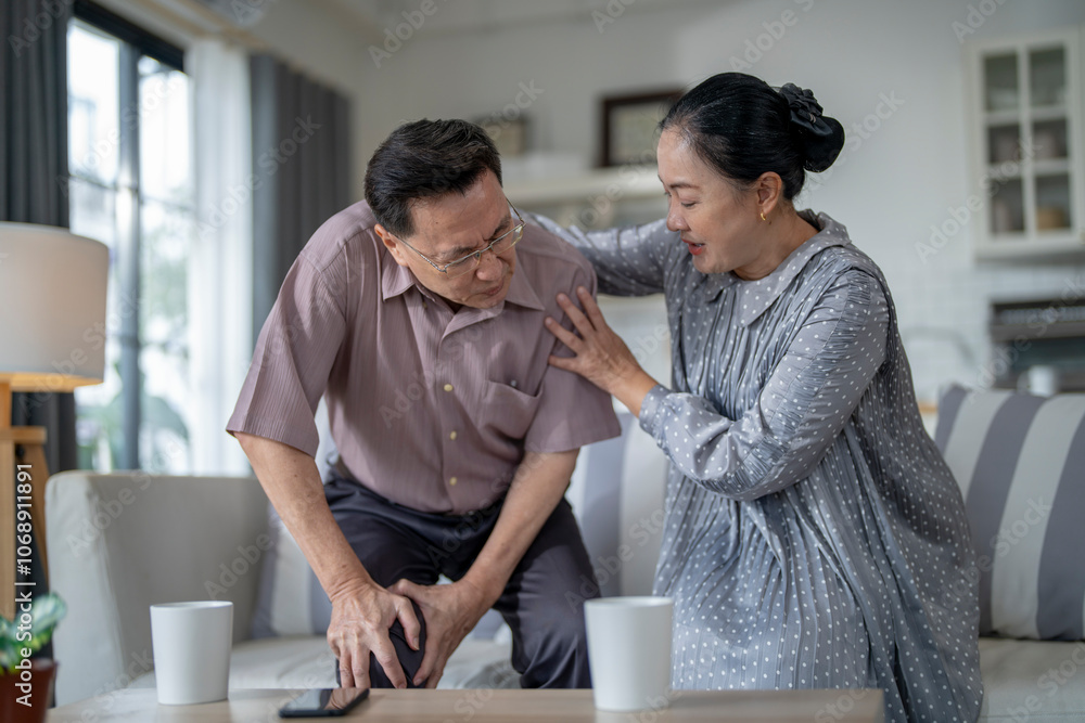 An elderly couple sits in their living room. The man holds his knee, appearing to be in discomfort, while his partner offers support. The image conveys care, concern, and the bond between the couple. An elderly couple sits in their living room. The man holds his knee, appearing to be in discomfort, while his partner offers support. The image conveys care, concern, and the bond between the couple.
