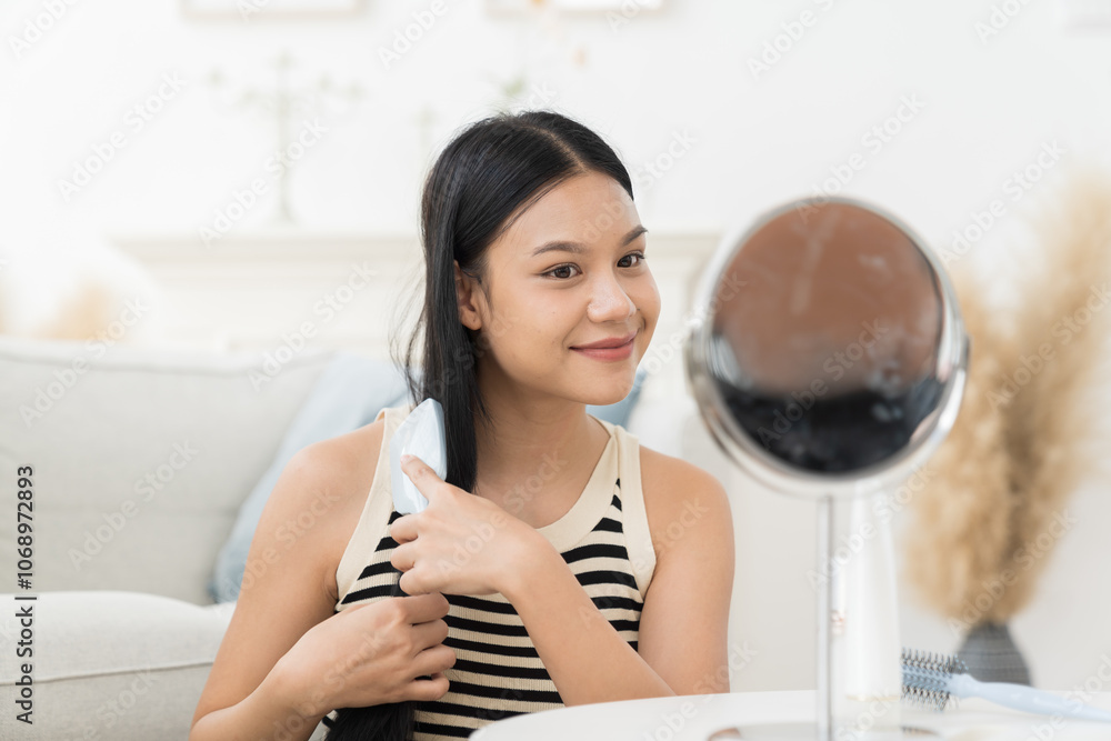 Cheerful young Asian pretty woman combing her beautiful hair with brush while sitting near mirror, looking at mirror with happy and smiling in living room Cheerful young Asian pretty woman combing her beautiful hair with brush while sitting near mirror, looking at mirror with happy and smiling in living room