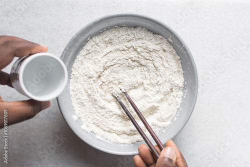 Overhead view of noodle dough being being mixed in a grey bowl, top view of flour and water being mixed with chopsticks to make noodles, process of making hand pulled noodles