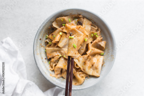 Overhead view of spicy wide noodles in chopsticks, top view of biangbiang mian noodle, spicy belt noodles with green onions and diced pork in a bowl