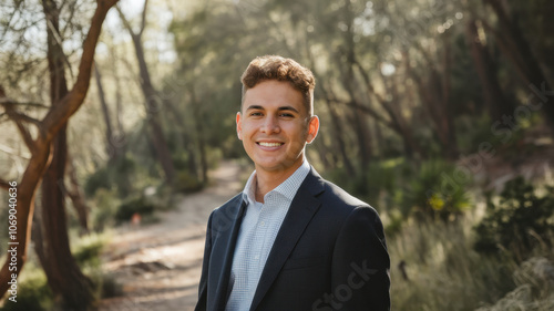 A young Hispanic man, approximately 20 years old, smiles confidently in a natural setting, featuring lush greenery and gentle sunlight filtering through the trees.