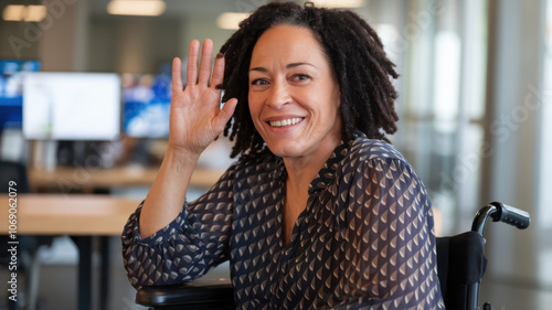 Smiling African American woman in a wheelchair waves hello in a modern office environment, showcasing a welcoming and friendly demeanor.