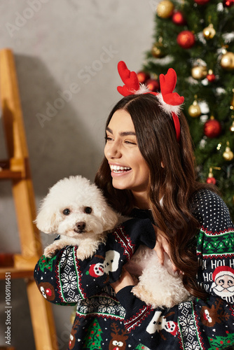 A joyful young woman in a cozy sweater celebrates Christmas with her Bichon Frise at home photo