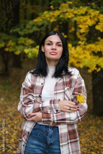 Young woman with dark hair in a plaid shirt holding a yellow leaf in an autumn forest. Captures the beauty of fall, nature, and outdoor lifestyle. Perfect for seasonal or nature-themed visuals.