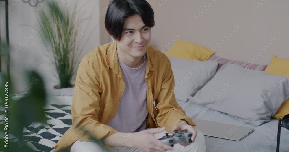 Joyful Asian man playing online video game using wireless controller and earphones sitting on bed having fun at home in leisure time. Gaming and people concept. Joyful Asian man playing online video game using wireless controller and earphones sitting on bed having fun at home in leisure time. Gaming and people concept.