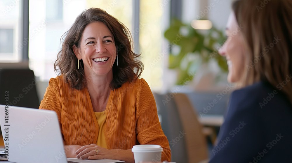 Collaborative office joy: A scene featuring two female professionals laughing and conversing near a desk with a laptop and coffee cup, exemplifying a collaborative and welcoming of Collaborative office joy: A scene featuring two female professionals laughing and conversing near a desk with a laptop and coffee cup, exemplifying a collaborative and welcoming of