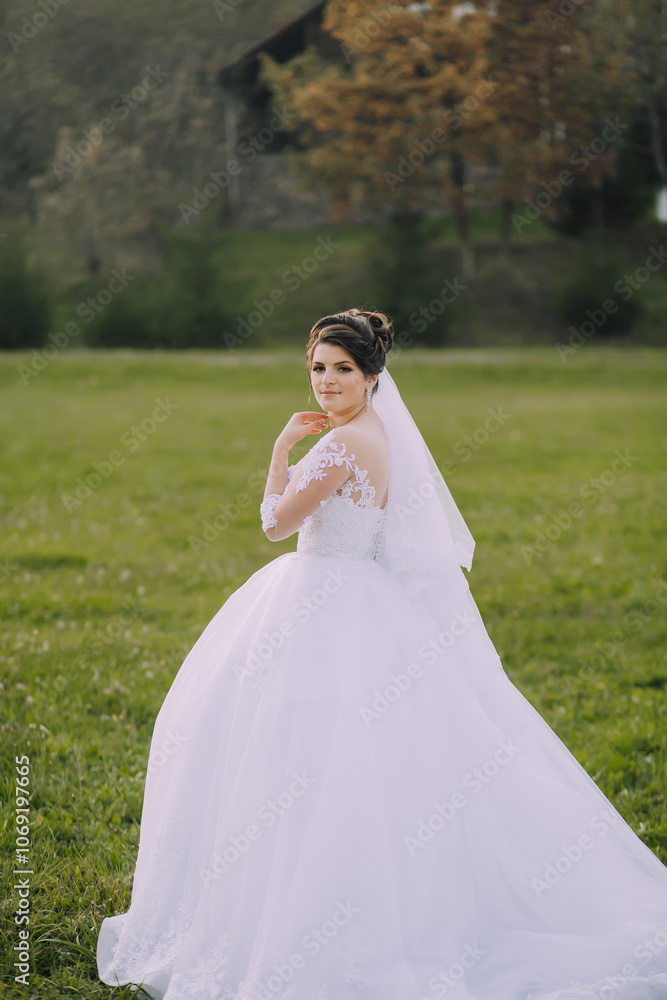 A woman in a white wedding dress stands in a field. She is wearing a veil and has her hands on her hips