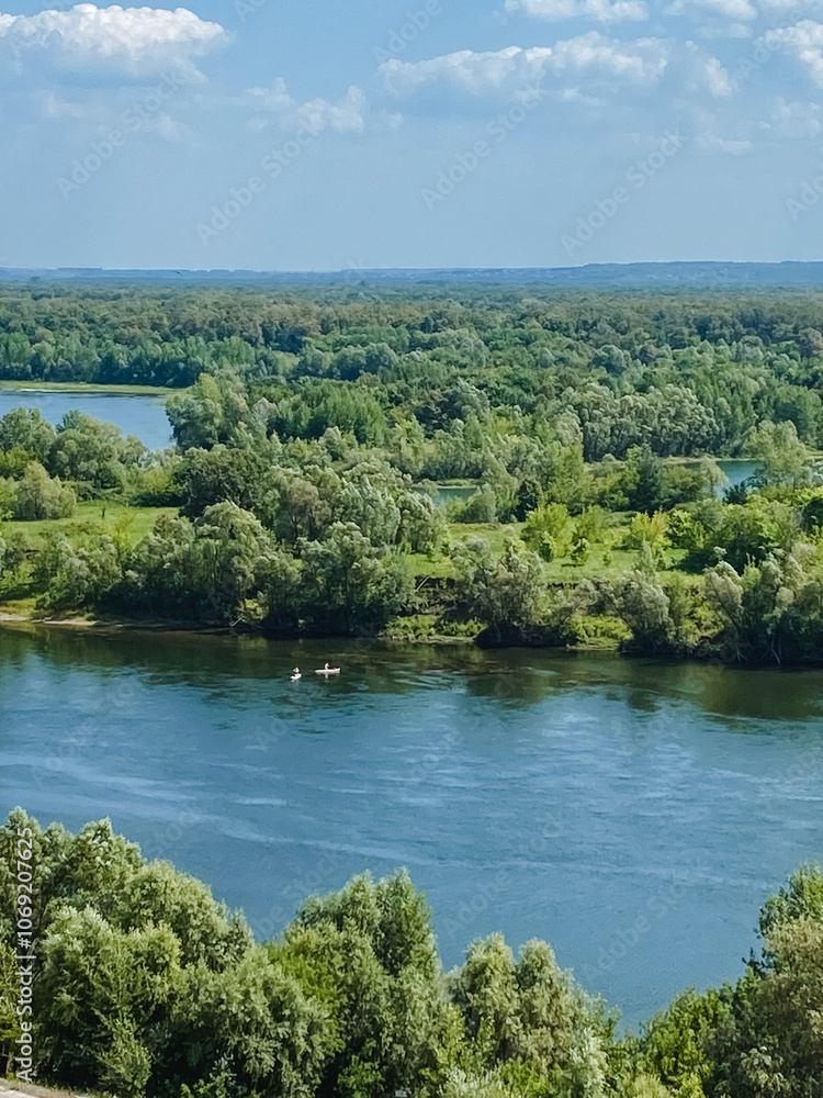 Naklejka premium A large and serene body of water, glistening under the sun, is beautifully surrounded by towering trees, all basking in the delightful warmth of a sunny day on a picturesque landscape