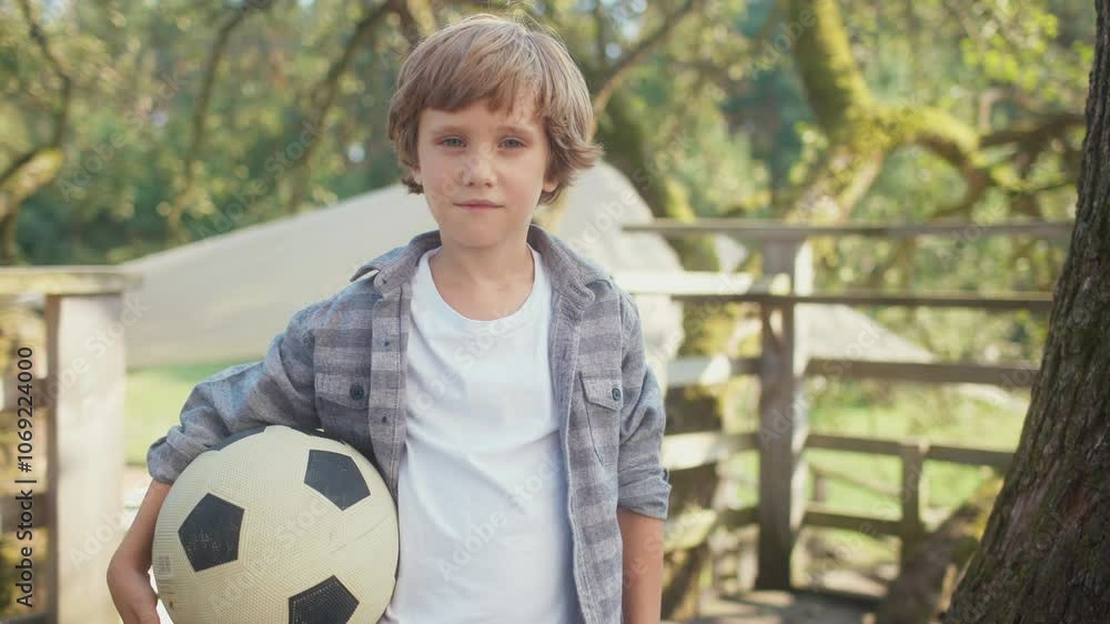Front camera view of handsome little boy standing in green garden. Holding big soccer ball with one hand. Wearing checkered shirt and white t-shirt. Spending free time actively. Looking at camera. Front camera view of handsome little boy standing in green garden. Holding big soccer ball with one hand. Wearing checkered shirt and white t-shirt. Spending free time actively. Looking at camera.