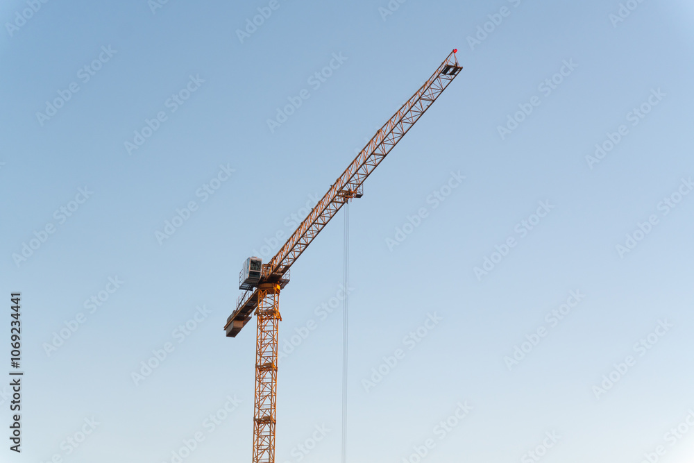 A yellow tower crane against a cloudless sky. The bright color of the crane stands out in the minimalist composition against the blue background. A yellow tower crane against a cloudless sky. The bright color of the crane stands out in the minimalist composition against the blue background.