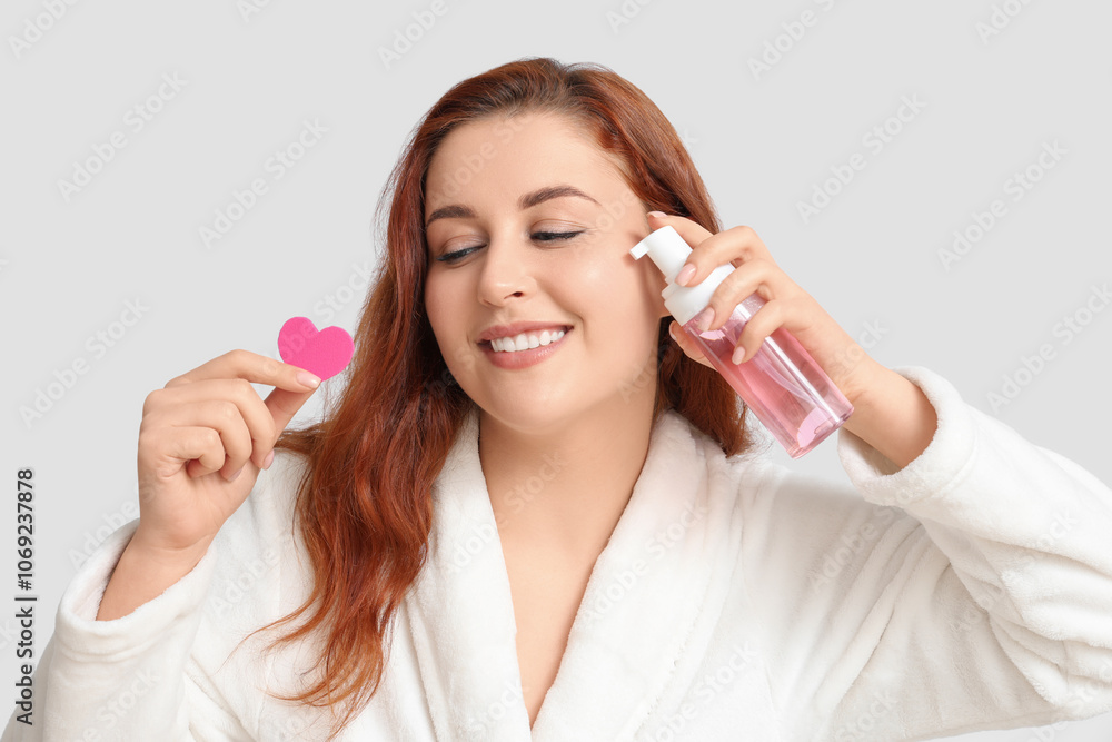 Beautiful young woman holding heart-shaped makeup sponge and bottle of cosmetic product on white background Beautiful young woman holding heart-shaped makeup sponge and bottle of cosmetic product on white background