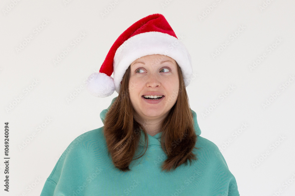 Surprised young brunette Santa woman in Christmas hat keeping smile looking aside isolated on white background, studio portrait. Happy New Year celebration merry holiday concept