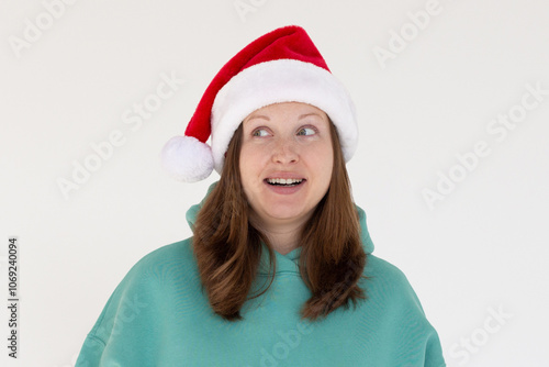 Surprised young brunette Santa woman in Christmas hat keeping smile looking aside isolated on white background, studio portrait. Happy New Year celebration merry holiday concept