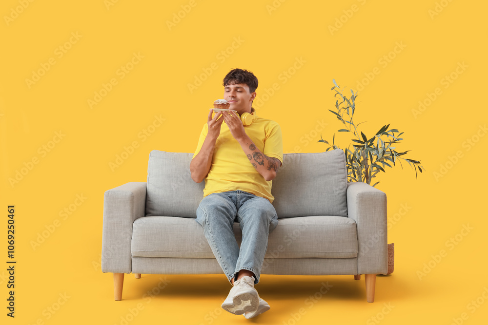 Obraz premium Young man with tasty muffin sitting on sofa against yellow background