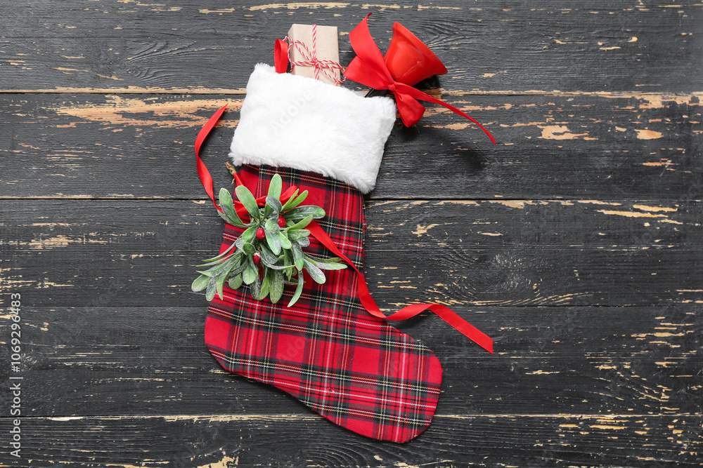 Naklejka premium Christmas sock with mistletoe twig, bell and gift box on black wooden background. Top view