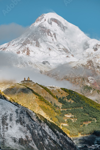 Gergeti Trinity Church perched on a hill with the snow-covered Mount Kazbek towering in the background, under a vibrant blue sky in Georgia. photo