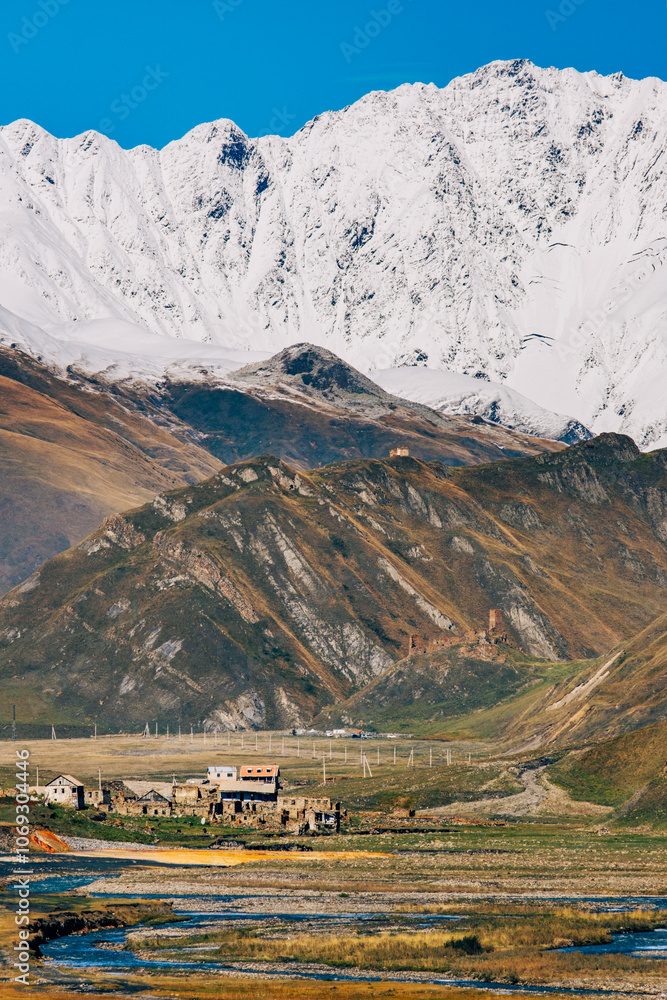 Obraz premium Alpine landscape with the village of Ketrisi, winding rivers, snowy peaks, and a serene trail under a clear blue sky in the Truso Gorge, near Kazbegi, Georgia