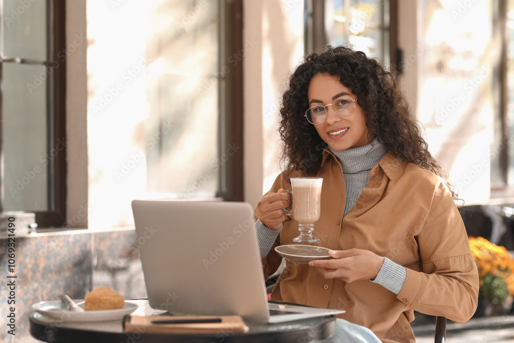 Young African-American author with cup of coffee and laptop sitting at table in street cafe Young African-American author with cup of coffee and laptop sitting at table in street cafe