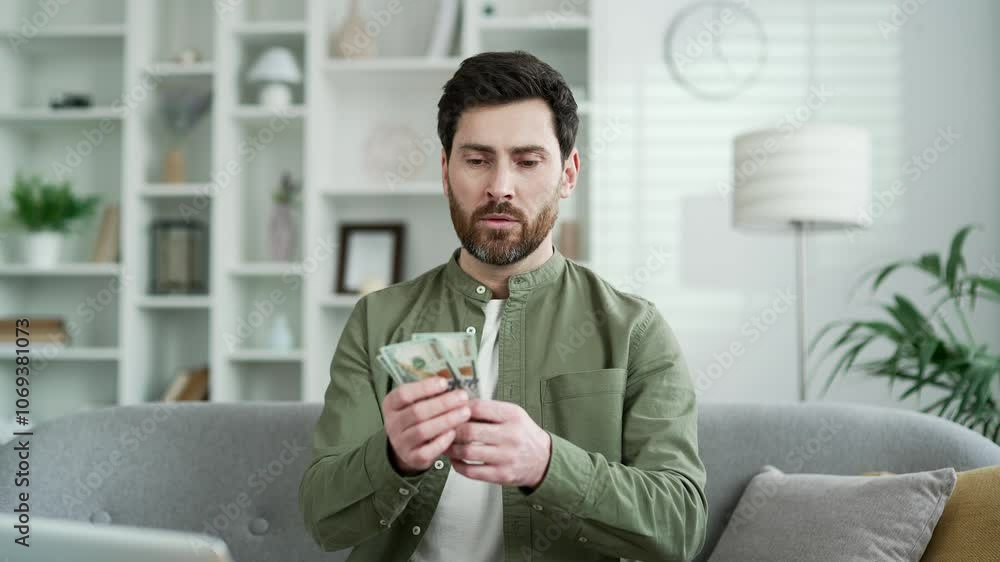 Handsome man counting dollar bills while sitting on the sofa in the living room at home. Thoughtful bearded employee holding cash in hands, calculates his earnings, not satisfied with his salary Handsome man counting dollar bills while sitting on the sofa in the living room at home. Thoughtful bearded employee holding cash in hands, calculates his earnings, not satisfied with his salary