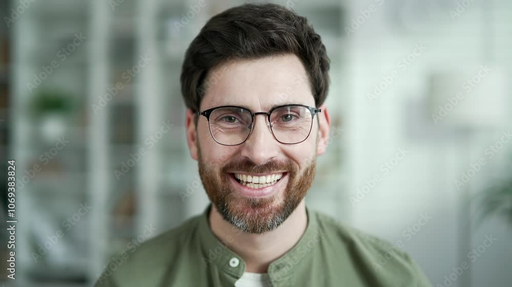 Portrait of happy handsome man sitting in living room at home. Head shot of confident smiling bearded businessman in shirt. Positive mature male in glasses poses looking at camera indoors. Close up Portrait of happy handsome man sitting in living room at home. Head shot of confident smiling bearded businessman in shirt. Positive mature male in glasses poses looking at camera indoors. Close up