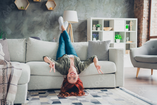 Full length photo of lovely young ginger girl lying upside down v-sign dressed khaki garment spacious house indoors room