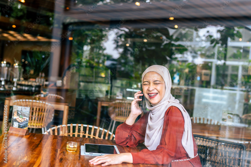 A joyful woman in a hijab is laughing while talking on her smartphone inside a cozy cafe. The image captures a warm, cheerful atmosphere, reflecting connection and happiness in a modern setting. A joyful woman in a hijab is laughing while talking on her smartphone inside a cozy cafe. The image captures a warm, cheerful atmosphere, reflecting connection and happiness in a modern setting.