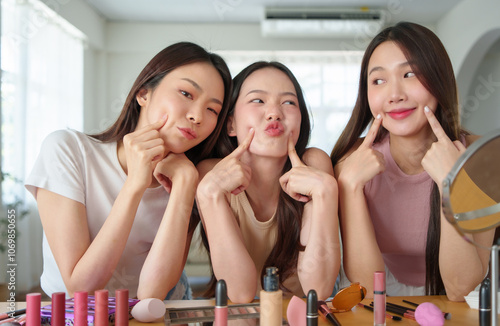 Three young Asian women having fun with makeup, Happy Korean girls making playful faces in a beauty setup with cosmetics on table, expressing joy and friendship.