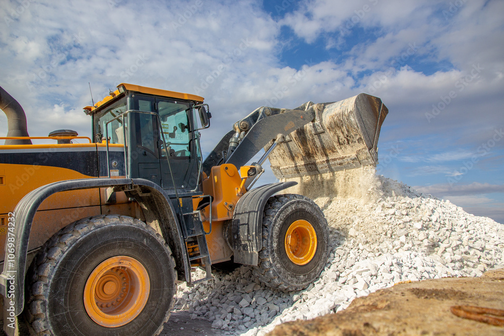 a yellow bucket truck dumps white marble chips onto an open warehouse at the wharf. A bucket loader forms a stack a yellow bucket truck dumps white marble chips onto an open warehouse at the wharf. A bucket loader forms a stack