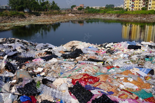 A large pile of discarded textiles and waste materials accumulating near a body of water likely representing environmental pollution