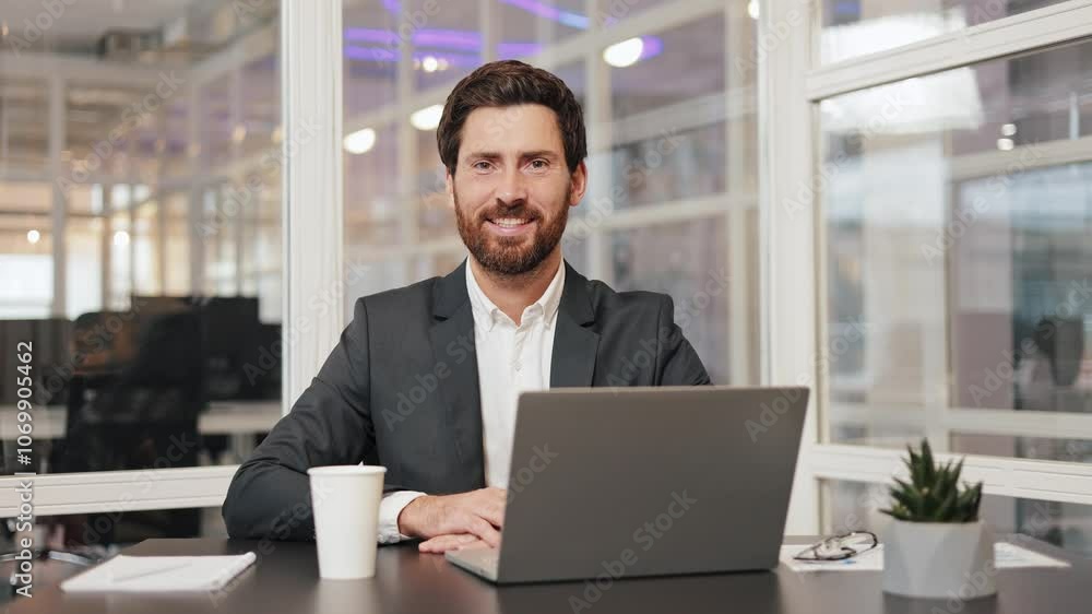 Friendly positive businessman in a suit sits at desk in office, working on laptop. Professional financial advisor, executive leader, manager, male lawyer entrepreneur looks up and smiles at camera. Friendly positive businessman in a suit sits at desk in office, working on laptop. Professional financial advisor, executive leader, manager, male lawyer entrepreneur looks up and smiles at camera.