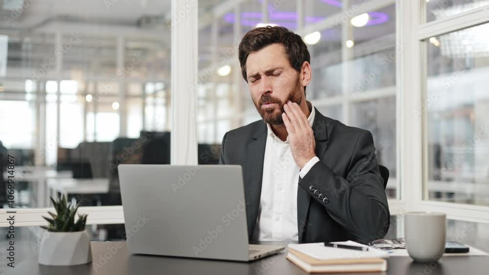 Businessman in his modern office working on a laptop experiences a sudden toothache or jaw pain. He is rubbing his jaw and face with concern. The modern office space is visible in the background. Businessman in his modern office working on a laptop experiences a sudden toothache or jaw pain. He is rubbing his jaw and face with concern. The modern office space is visible in the background.