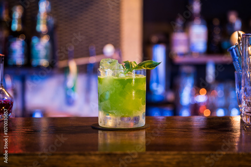 A bright green cocktail sits on a wooden bar, garnished with basil leaves and ice cubes. The background is blurred with soft, colorful bar lights.