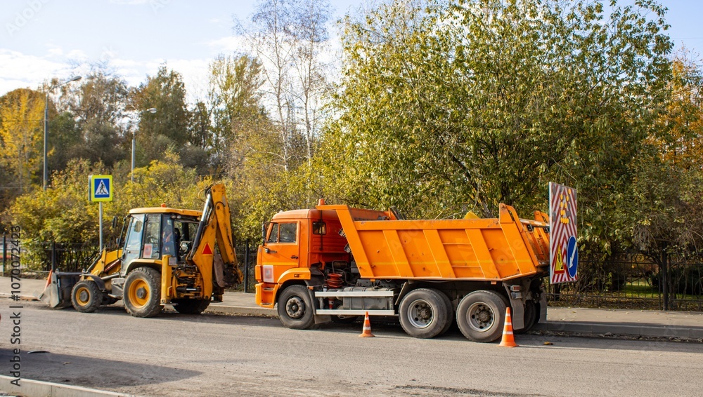 A bulldozer and a large truck are ready to begin road work. Road signs indicating a detour around the obstacle are installed on the dump truck body. A bulldozer and a large truck are ready to begin road work. Road signs indicating a detour around the obstacle are installed on the dump truck body.