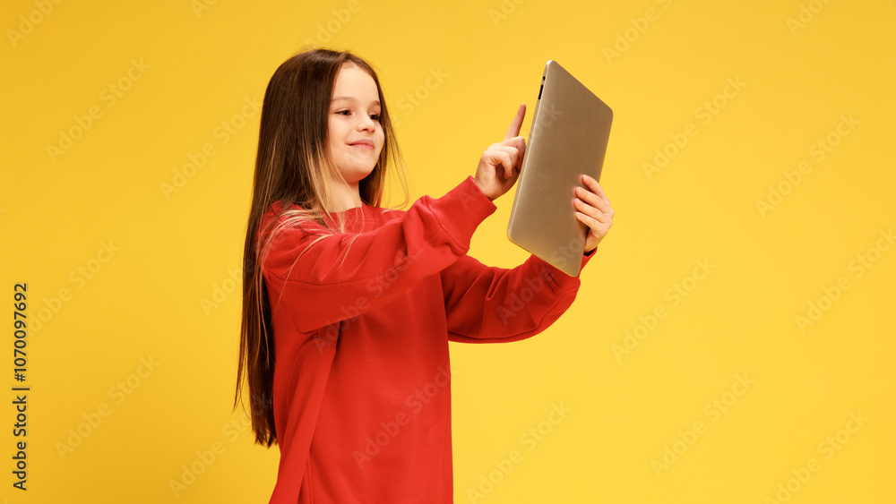 Girl in red sweater pointing at her tablet, as she choose something, and smiling against yellow studio background. Concept of childhood, lifestyle, expressions, trendy. Ad Girl in red sweater pointing at her tablet, as she choose something, and smiling against yellow studio background. Concept of childhood, lifestyle, expressions, trendy. Ad