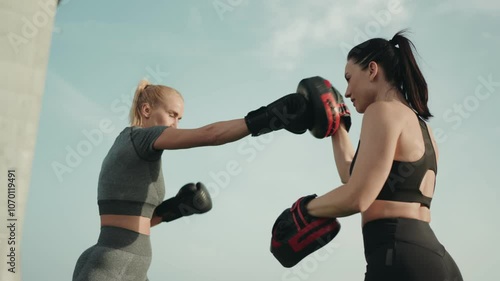 Two sportive women training together, practicing boxing techniques outdoors under a clear sky, showcasing strength and fitness.