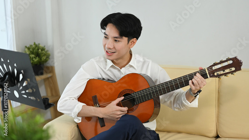 Smiling young man playing acoustic guitar with sheet music stand in the living room photo