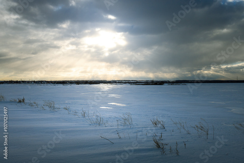 A snowy field with a cloudy sky and a sun shining through the clouds