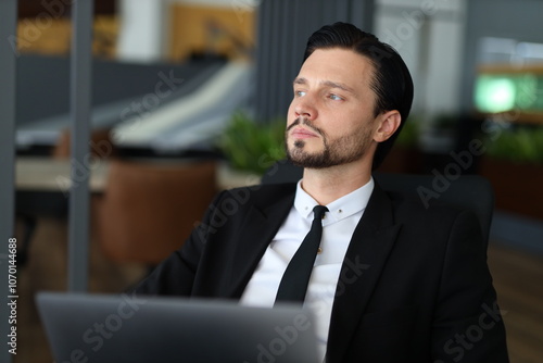 A man in a suit and tie is sitting at a desk with a laptop open in front of him. He is focused on his work, possibly preparing for a presentation or meeting