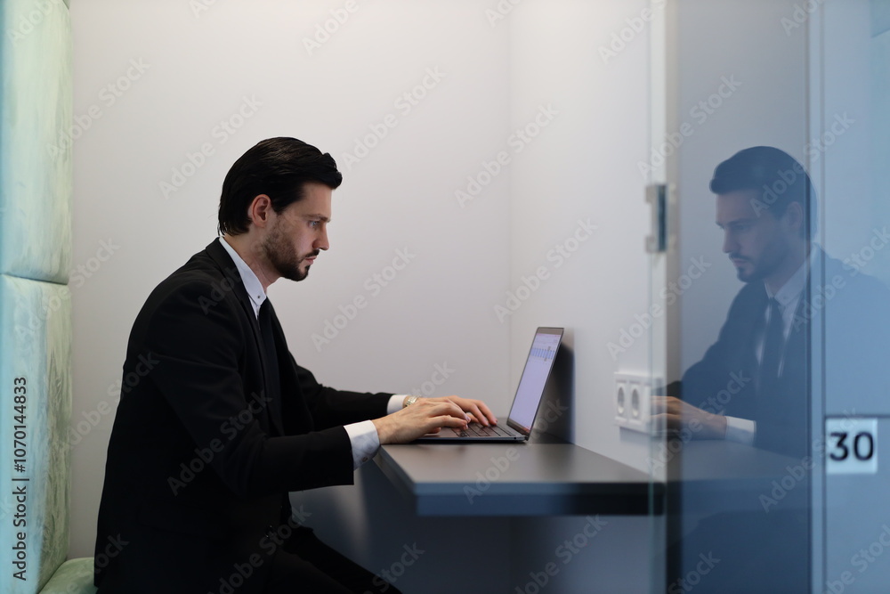 A man in a suit is typing on a laptop in a cubicle. The cubicle is a small, enclosed space with a glass door. The man is focused on his work, and the cubicle provides a sense of privacy A man in a suit is typing on a laptop in a cubicle. The cubicle is a small, enclosed space with a glass door. The man is focused on his work, and the cubicle provides a sense of privacy