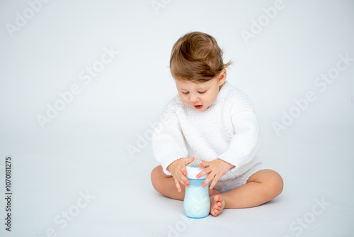 A baby boy with a bottle of milk on a white isolated background with a place for text is sitting in white knitted clothes. A small happy child drinks milk, space for text