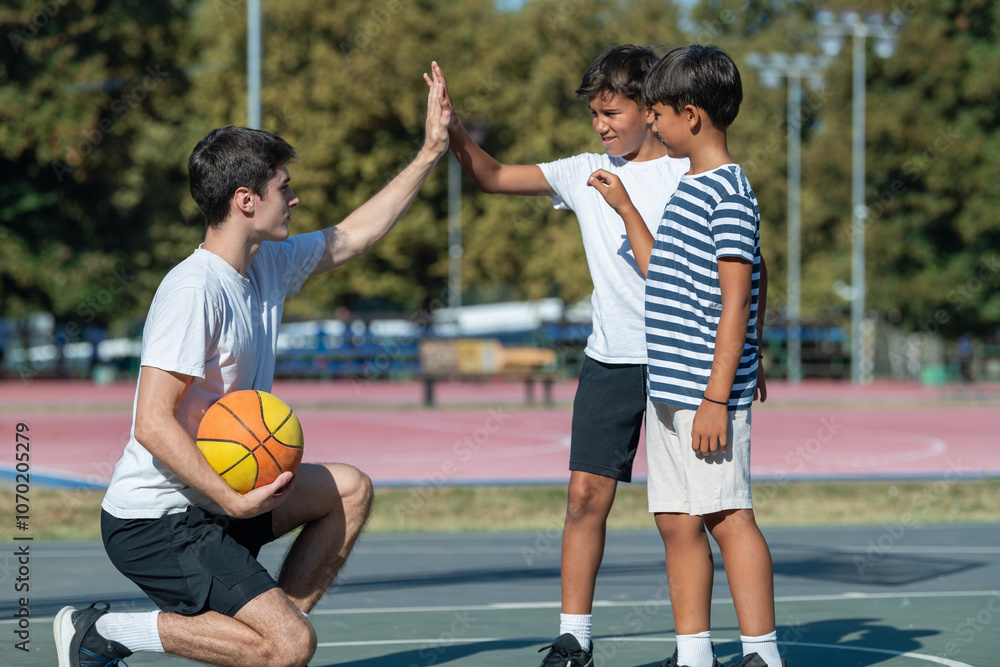 Obraz premium Coach and two young boys exchange a high five during outdoor basketball practice, capturing a moment of teamwork, encouragement, and active participation in youth sports