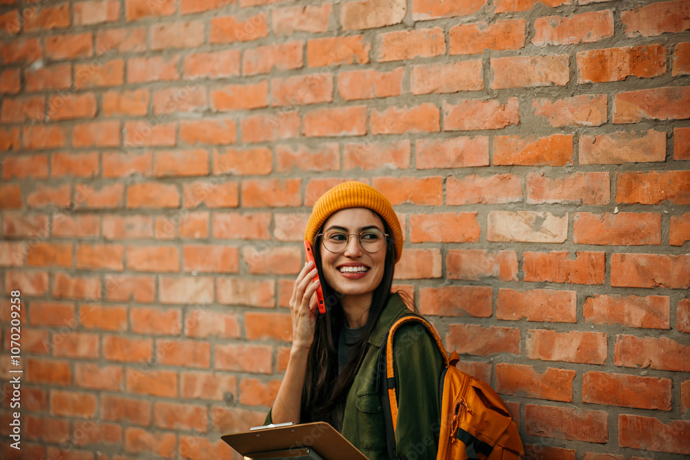 Naklejka premium Joyful academic female with stylish glasses carrying books and backpack, showcasing a trendy educational look against a brick background. Copy space