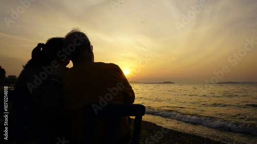 Fotografie Silhouette of an elderly couple sitting close together on the beach, watching a peaceful sunset over the sea, evoking feelings of companionship, love, and tranquility